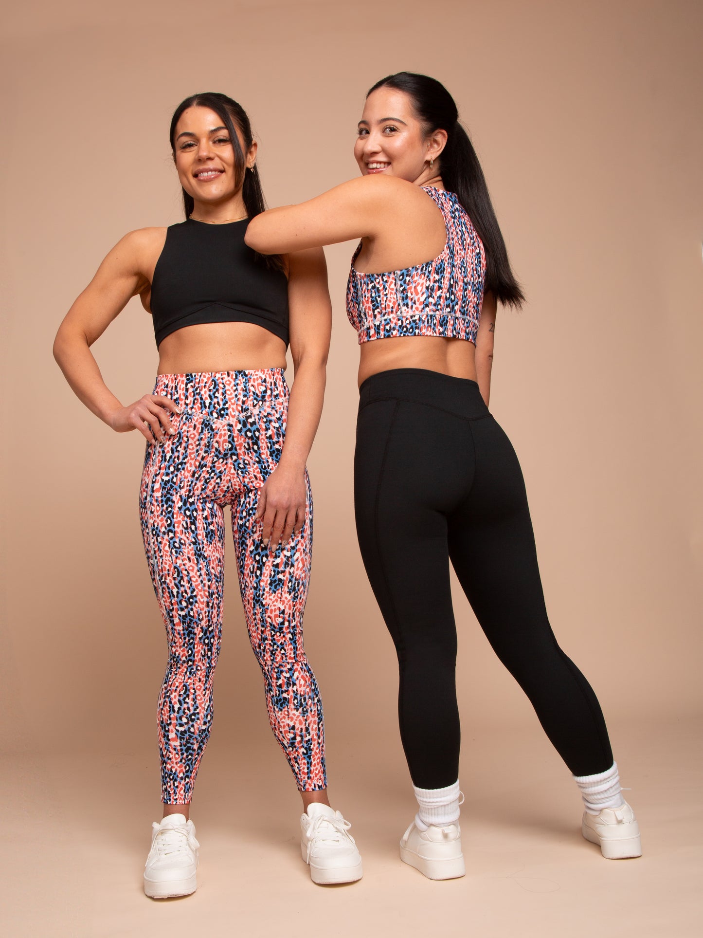 Two women in athleisure pose against a beige background. One smiles and touches her friend’s shoulder while wearing DEWEY’s IRIS High Waist Leggings (Printed, Extra Petite) with a coordinating crop top.