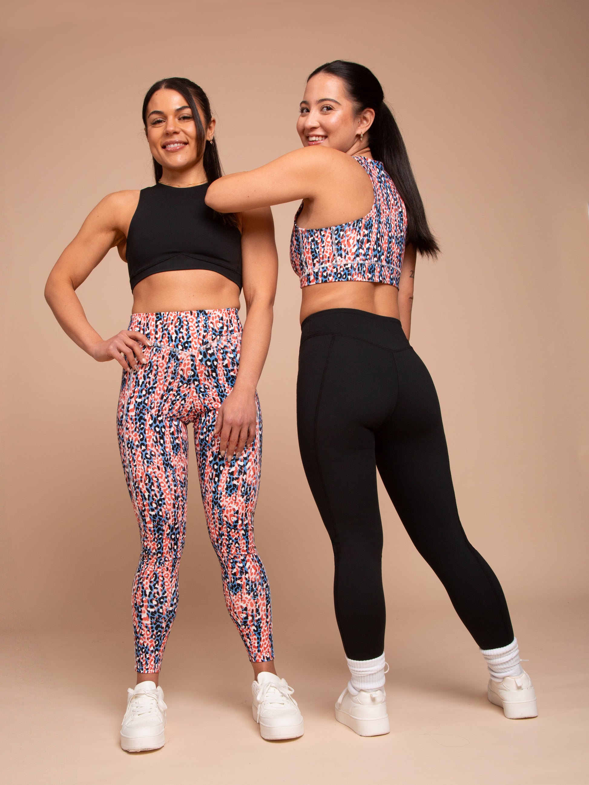 Two women in athleisure pose against a beige background. One smiles and touches her friend’s shoulder while wearing DEWEY’s IRIS High Waist Leggings (Printed, Extra Petite) with a coordinating crop top.