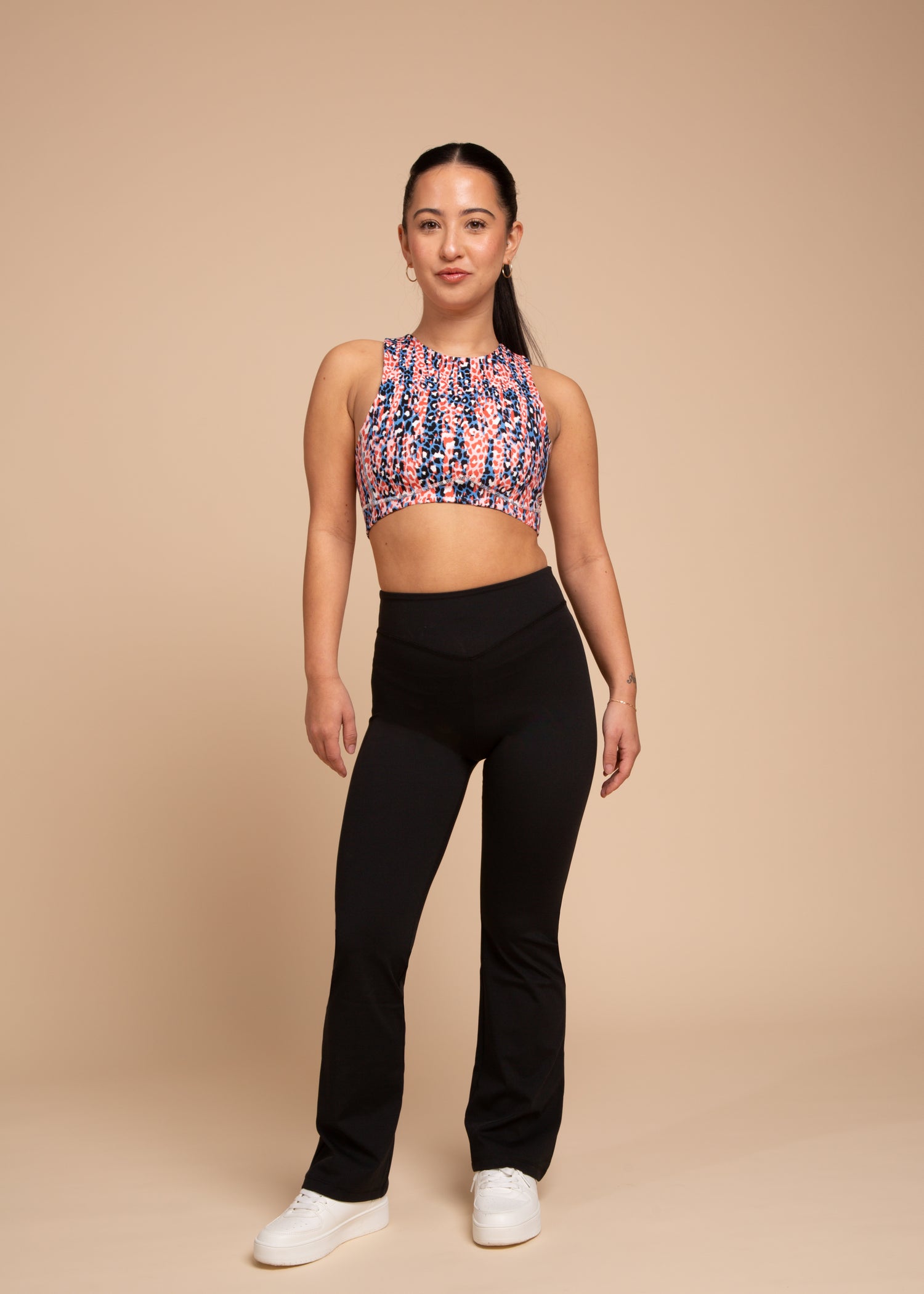 A woman with long dark hair tied back stands confidently against a beige background, wearing a colorful sleeveless crop top, white sneakers, and DEWEY MIRA Flared Leggings in Black (Extra Petite), with a relaxed expression.