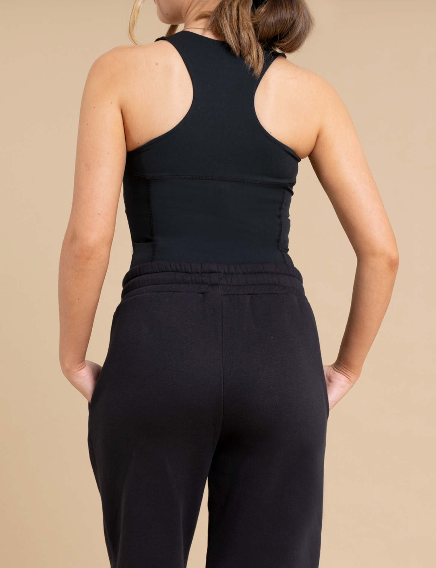 A person is seen from behind with hands in pockets, wearing the DEWEY SUMMER Racerback Vest Top in black (Extra Petite) and black sweatpants, standing against a beige background.