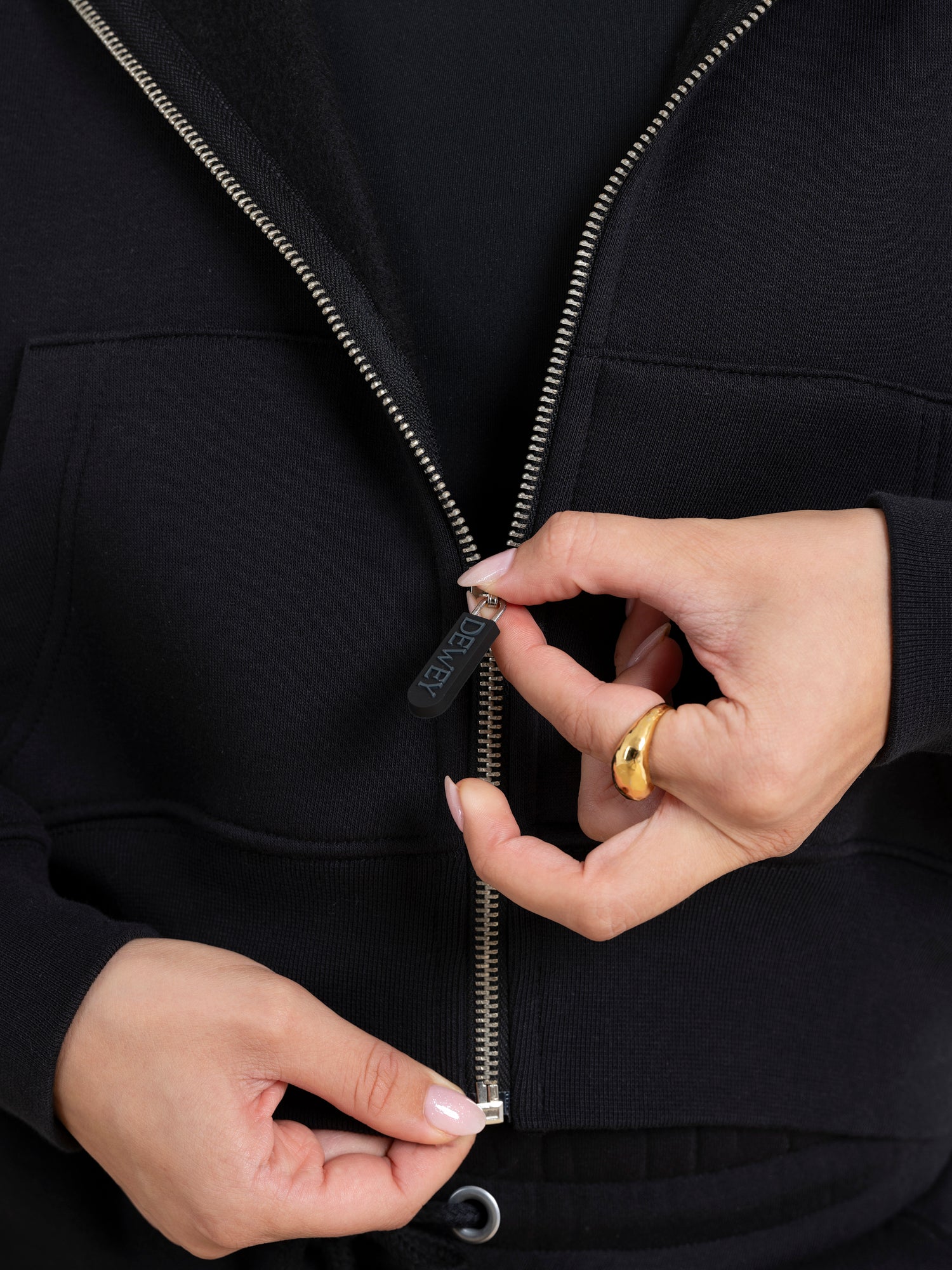 Close-up of a person zipping up the DEWEY TOBY Full Zip-Up Hoodie in Black (Extra Petite), one hand on the zipper pull, the other steadying the fabric, with a gold ring on their index finger.