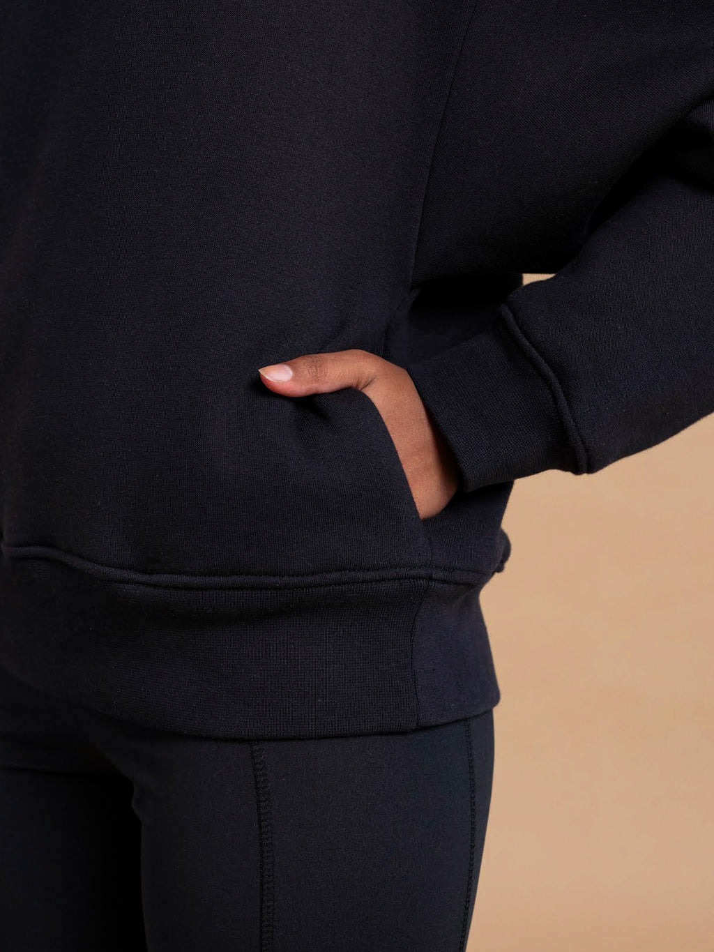 Close-up of a person wearing the DEWEY BAILEY Crewneck Sweatshirt Jumper in Black (Extra Petite) with black leggings, right hand in the pocket, against a beige background. The relaxed fit creates a cozy, effortless style.