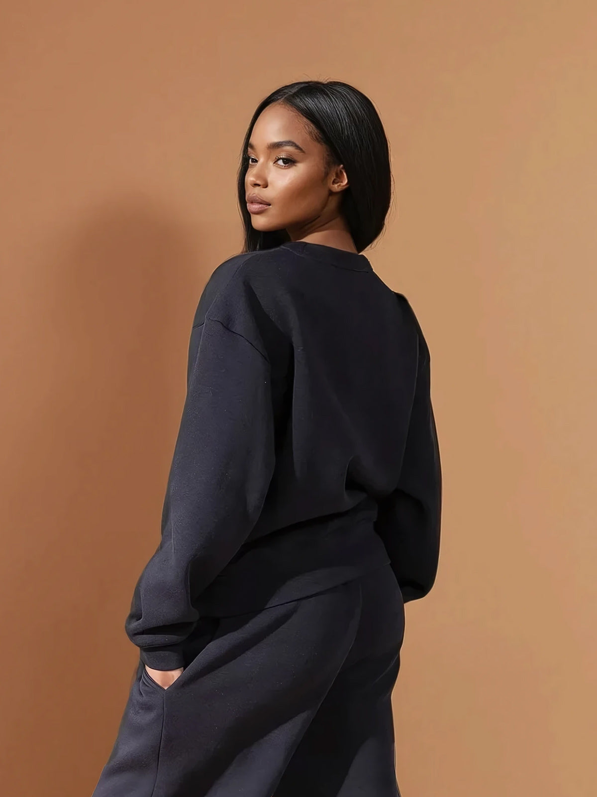 A woman with long, center-parted hair stands against a tan background, calmly looking over her shoulder. She wears a relaxed fit DEWEY BAILEY Crewneck Sweatshirt Jumper in Black (Extra Petite) paired with matching pants.