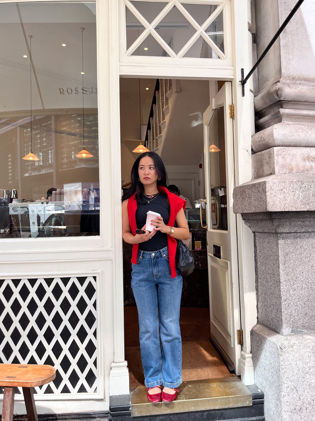 A woman in blue jeans and a DEWEY SUMMER Racerback Vest Top (Black, Extra Petite) with a red jacket on her shoulders and red shoes stands in a café doorway, holding a cup and glancing to the side.