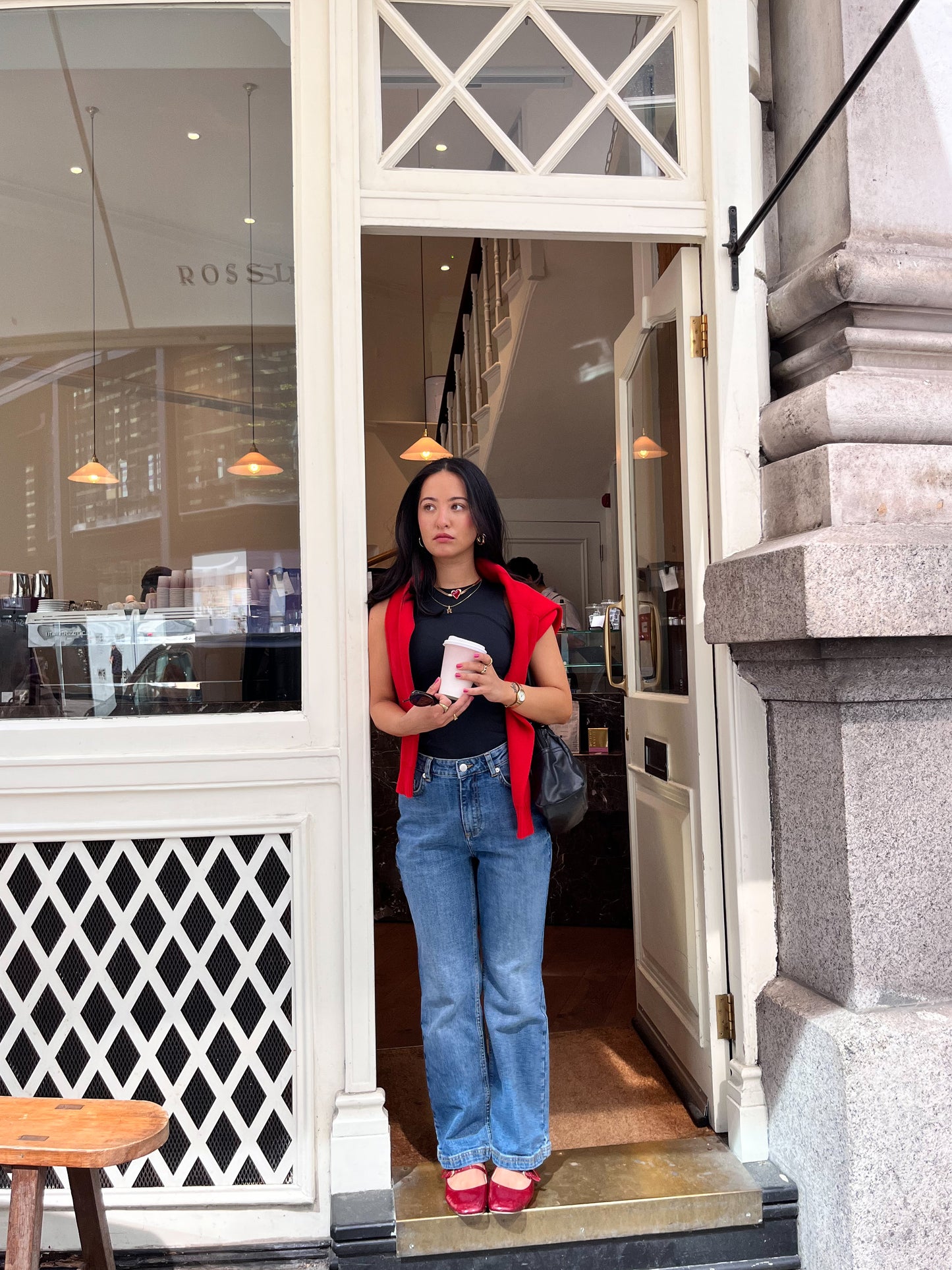 A woman in blue jeans and a DEWEY SUMMER Racerback Vest Top (Black, Extra Petite) with a red jacket on her shoulders and red shoes stands in a café doorway, holding a cup and glancing to the side.