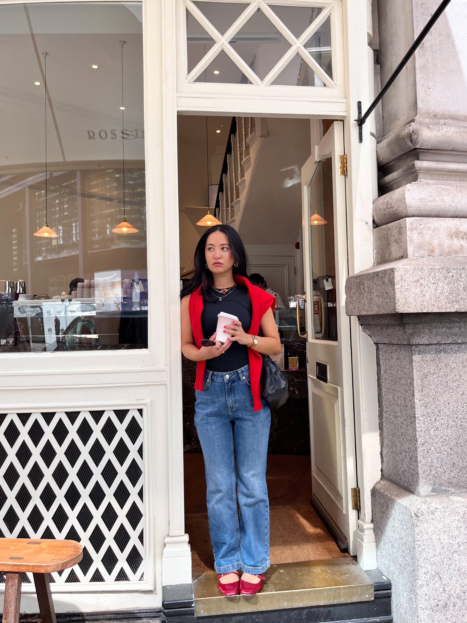A woman in blue jeans and a DEWEY SUMMER Racerback Vest Top (Black, Extra Petite) with a red jacket on her shoulders and red shoes stands in a café doorway, holding a cup and glancing to the side.