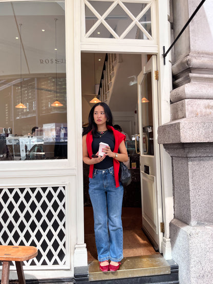 A woman in blue jeans and a DEWEY SUMMER Racerback Vest Top (Black, Extra Petite) with a red jacket on her shoulders and red shoes stands in a café doorway, holding a cup and glancing to the side.