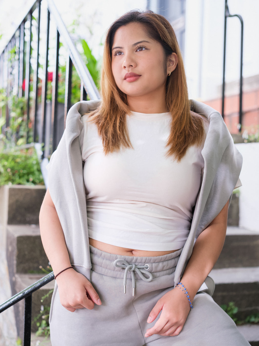 A woman with light brown hair sits outdoors on stairs, wearing a white shirt and DEWEY's BAILEY Crewneck Sweatshirt Jumper (Grey, Petite & Extra Petite) draped over her shoulders. She gazes thoughtfully away, with greenery and a railing behind her.
