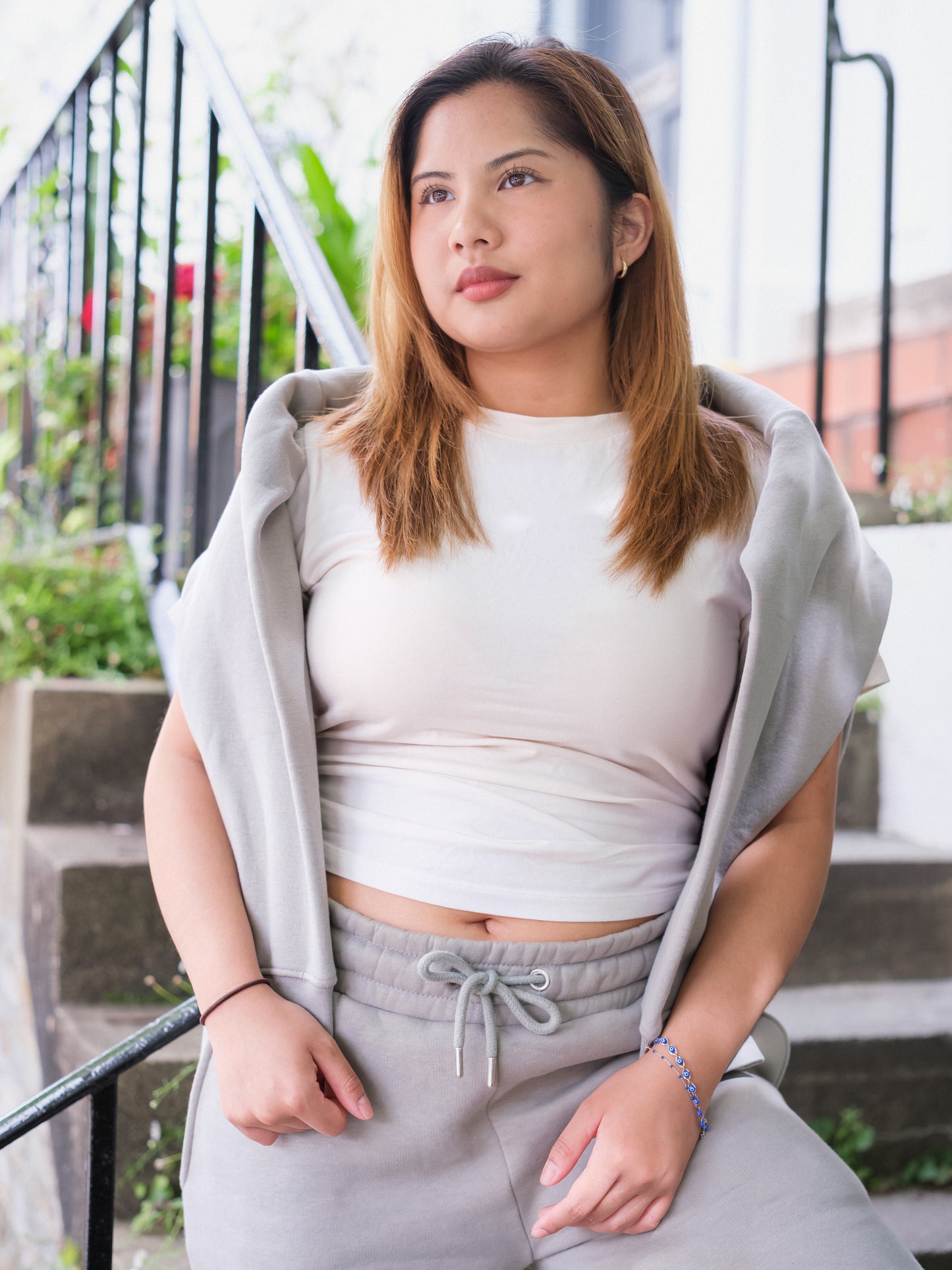 A woman with light brown hair sits outdoors on stairs, wearing a white shirt and DEWEY's BAILEY Crewneck Sweatshirt Jumper (Grey, Petite & Extra Petite) draped over her shoulders. She gazes thoughtfully away, with greenery and a railing behind her.
