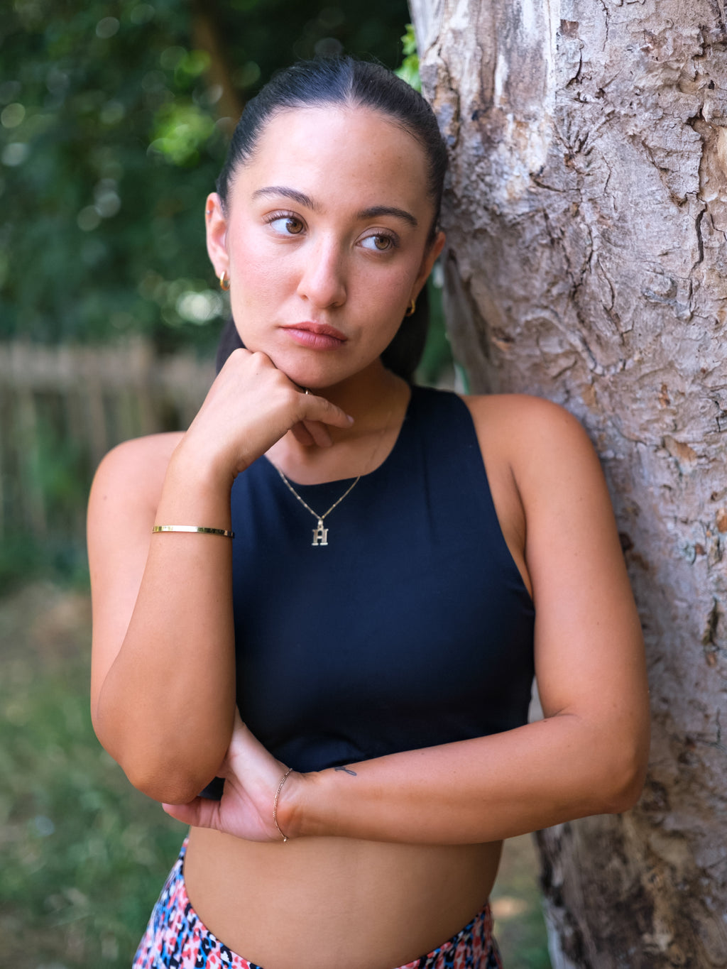 A woman with dark hair tied back and a thoughtful pose leans against a tree, showcasing the DEWEY HAZEL Cropped Vest Top in black (Extra Petite) amid lush greenery, highlighting her chic athleisure style.