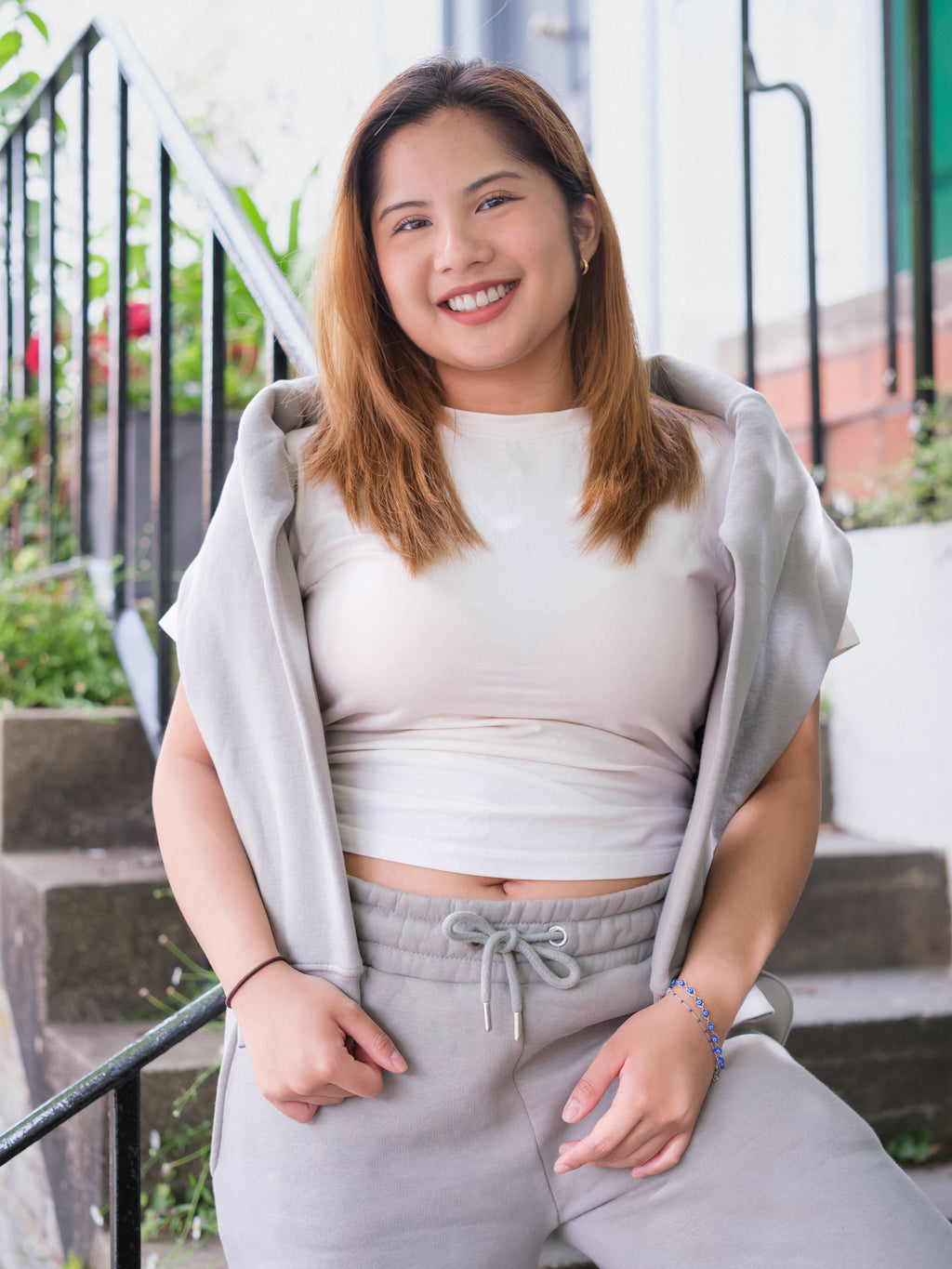 A young woman with light brown hair smiles while sitting on outdoor steps. She wears the DEWEY BAILEY Crewneck Sweatshirt Jumper (Grey, Petite & Extra Petite), with green plants and a railing in the background.