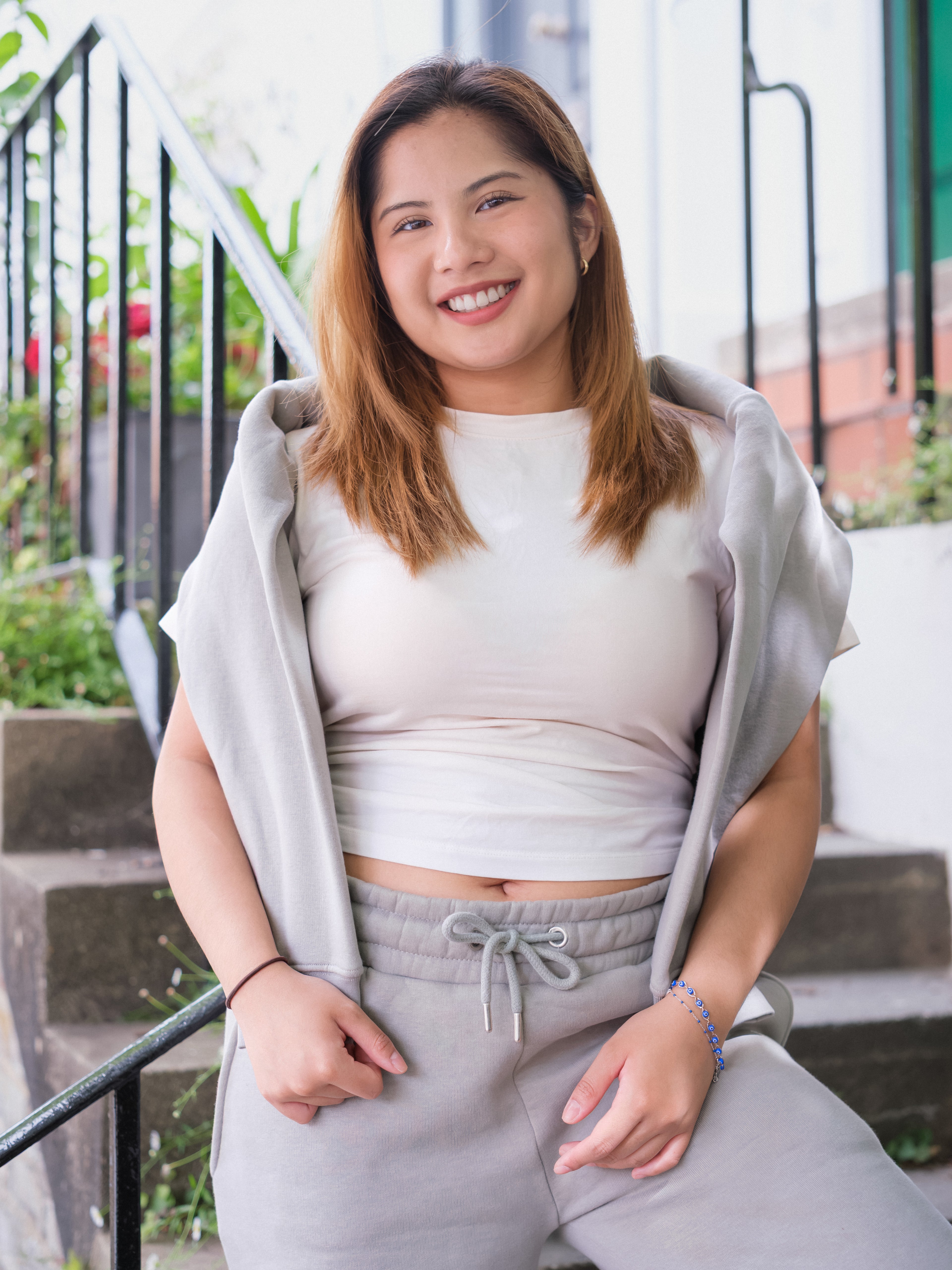 A young woman with light brown hair smiles while sitting on outdoor steps. She wears the DEWEY BAILEY Crewneck Sweatshirt Jumper (Grey, Petite & Extra Petite), with green plants and a railing in the background.