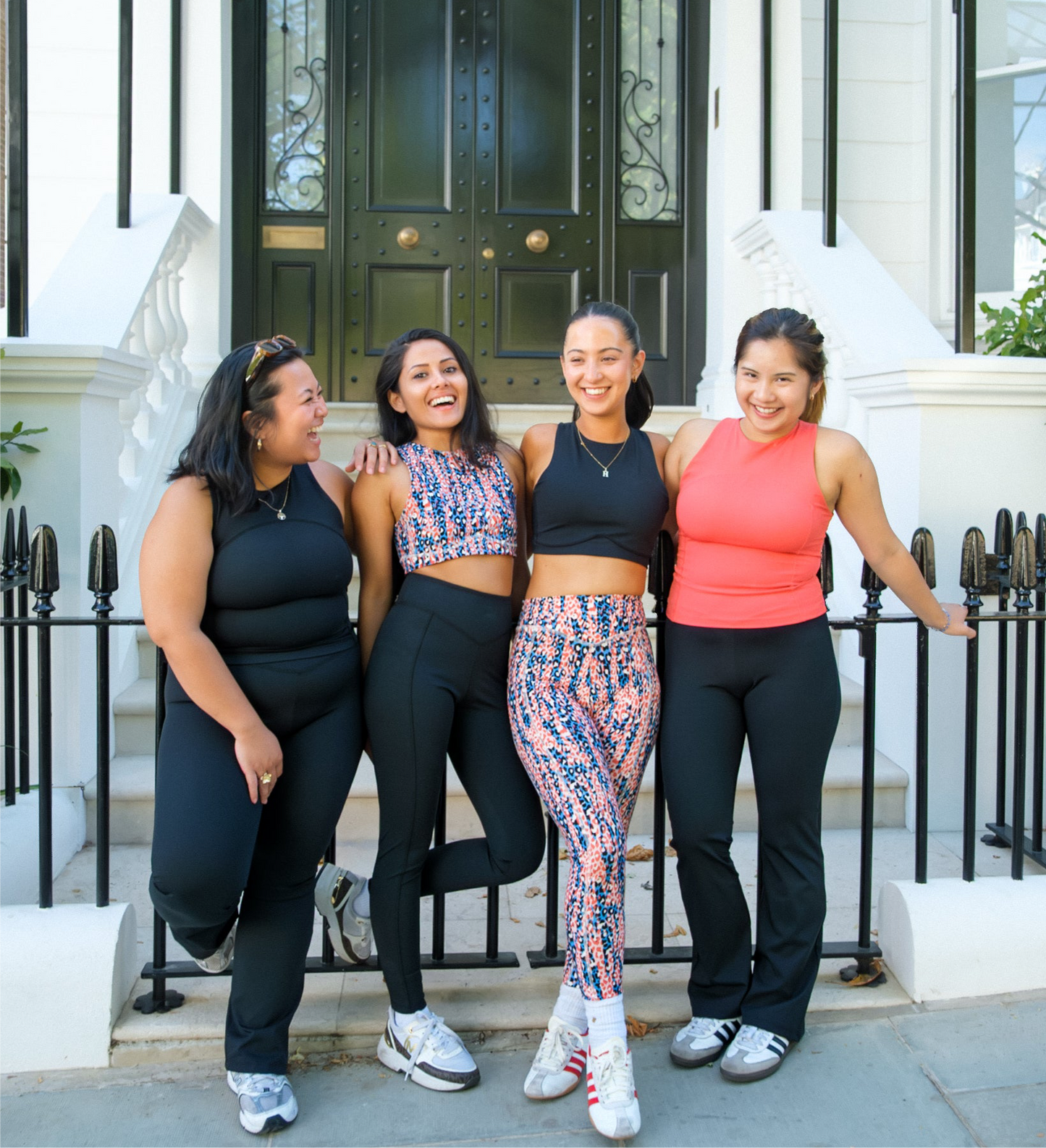 Four women in athleisure tops smile together outside. One wears the DEWEY SUMMER Racerback Vest Top in Coral Red (Extra Petite), ideal for active days out.