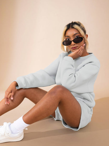 A woman wearing DEWEY’s RILEY Sweat Shorts in Grey (Extra Petite) sits on the floor against a beige background. She pairs them with a light gray sweatshirt, white sneakers and socks. Her hair is tied back, showing blonde highlights, and she wears sunglasses.