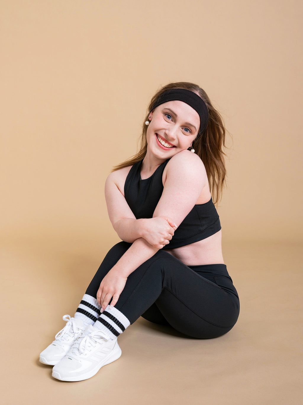 A smiling woman wears the DEWEY HAZEL Cropped Vest Top in Black (Extra Petite), paired with white sneakers, as she sits on the floor with her knees up and arms around them against a light beige background.