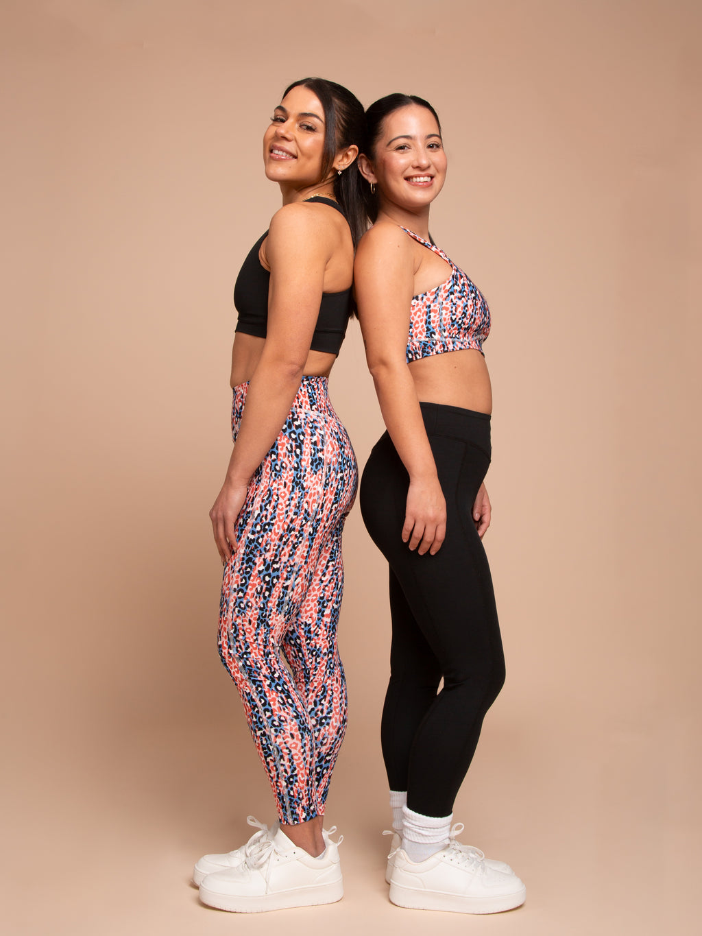 Two women, smiling back to back in white sneakers, model athleisure by DEWEY: one in the IRIS High Waist Leggings (Printed, Extra Petite) with a matching sports bra; the other in a patterned sports bra and black leggings against a beige backdrop.