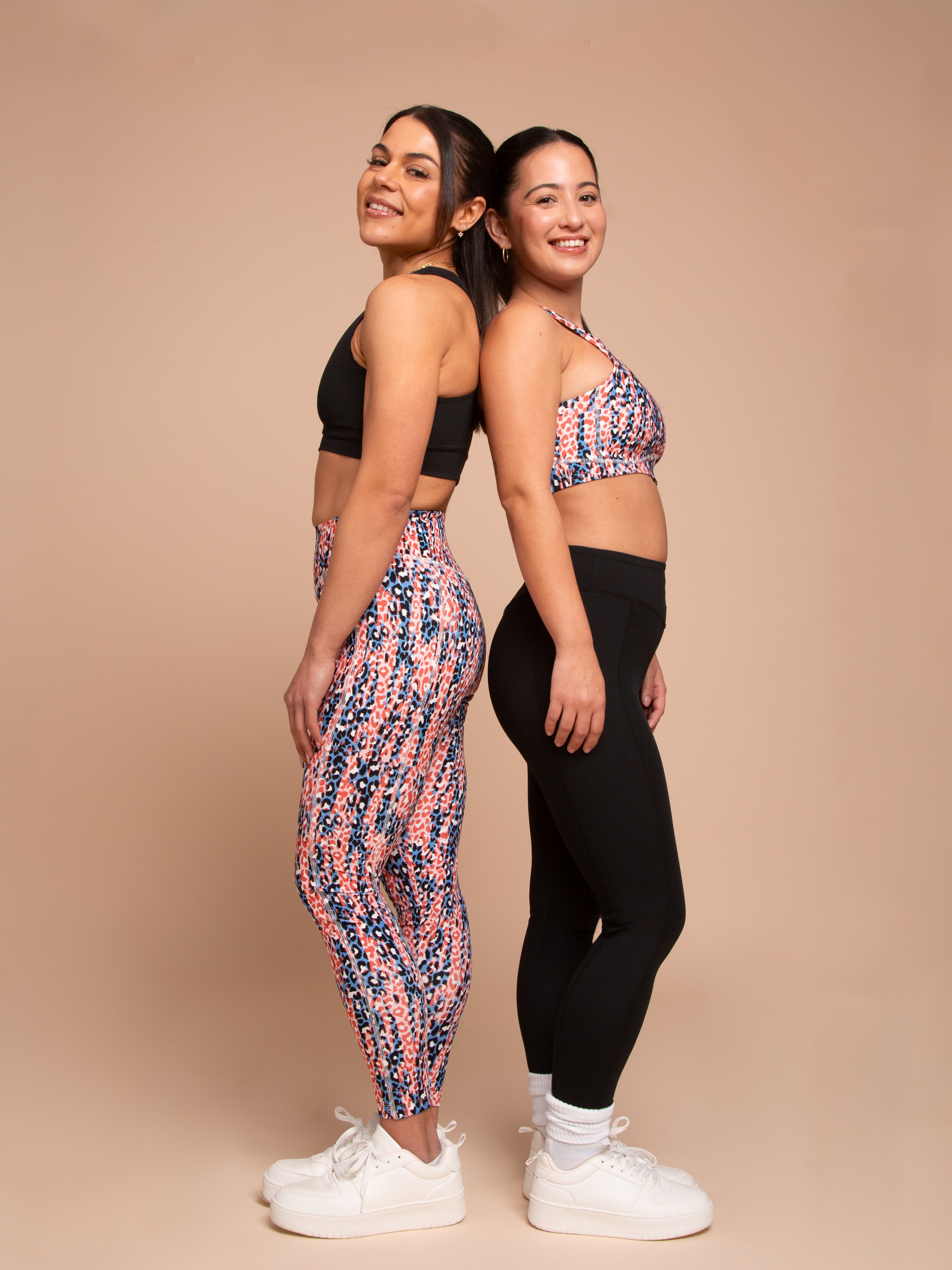 Two women, smiling back to back in white sneakers, model athleisure by DEWEY: one in the IRIS High Waist Leggings (Printed, Extra Petite) with a matching sports bra; the other in a patterned sports bra and black leggings against a beige backdrop.