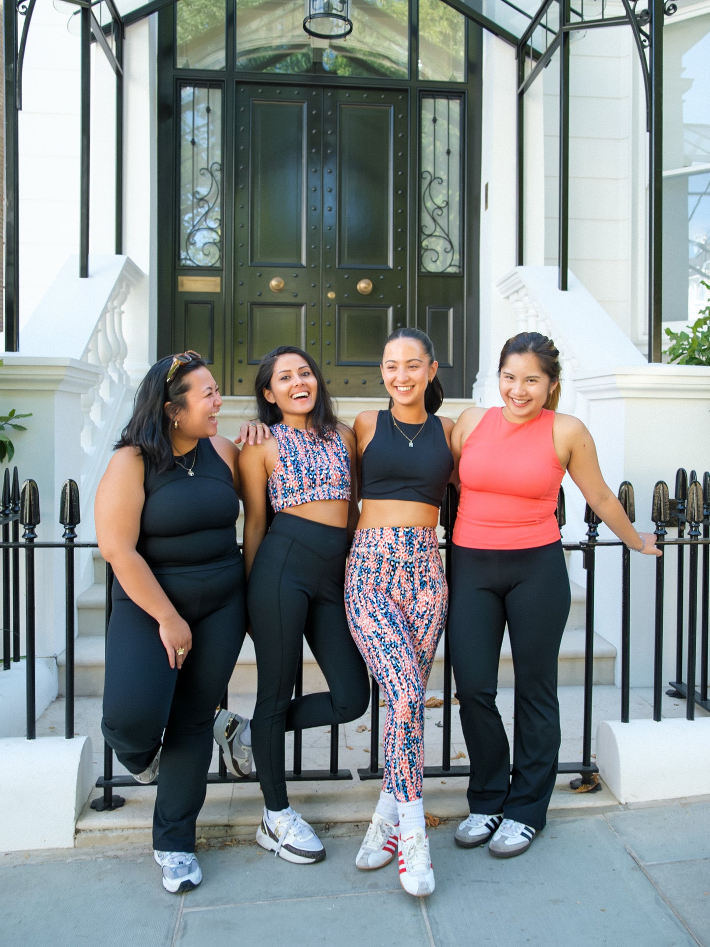 Four women in athletic wear, including one wearing the DEWEY SUMMER Racerback Vest Top (Black, Extra Petite), smile together in front of a white building with black doors, enjoying each other's company and posing for a photo.