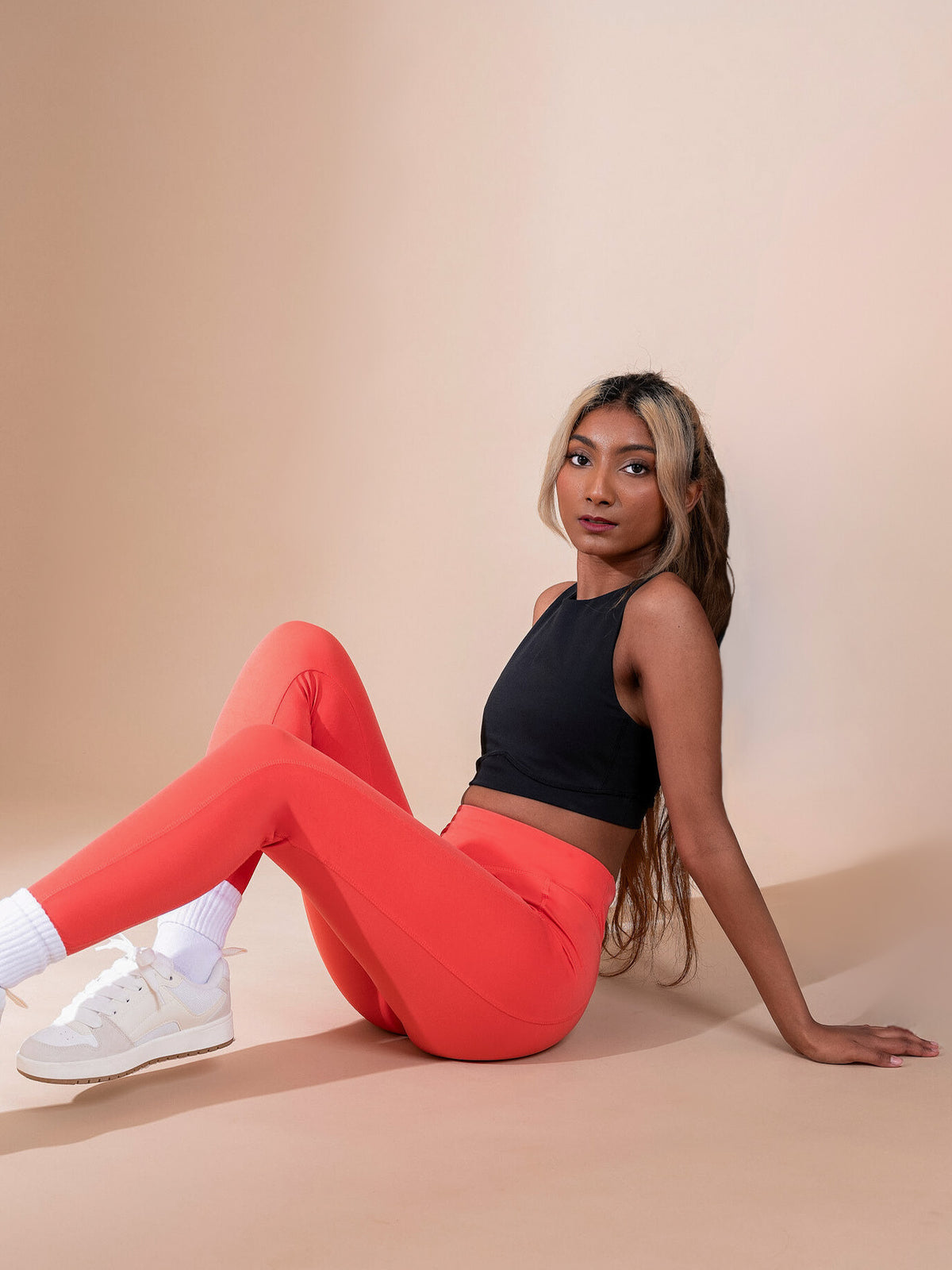 A woman with long hair sits on the floor against a beige background, wearing DEWEY’s IRIS High Waist Leggings in Coral Red (Extra Petite) paired with a black crop top, white socks, and white sneakers. She looks to the side with a relaxed expression.