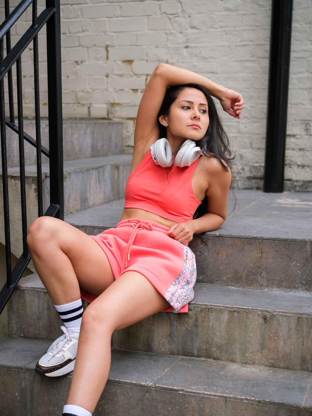 Wearing DEWEY's HAZEL Cropped Vest Top in Coral Red (Extra Petite), a woman sits relaxed on outdoor steps with headphones around her neck, pairing the athleisure look with white sneakers and striped socks.