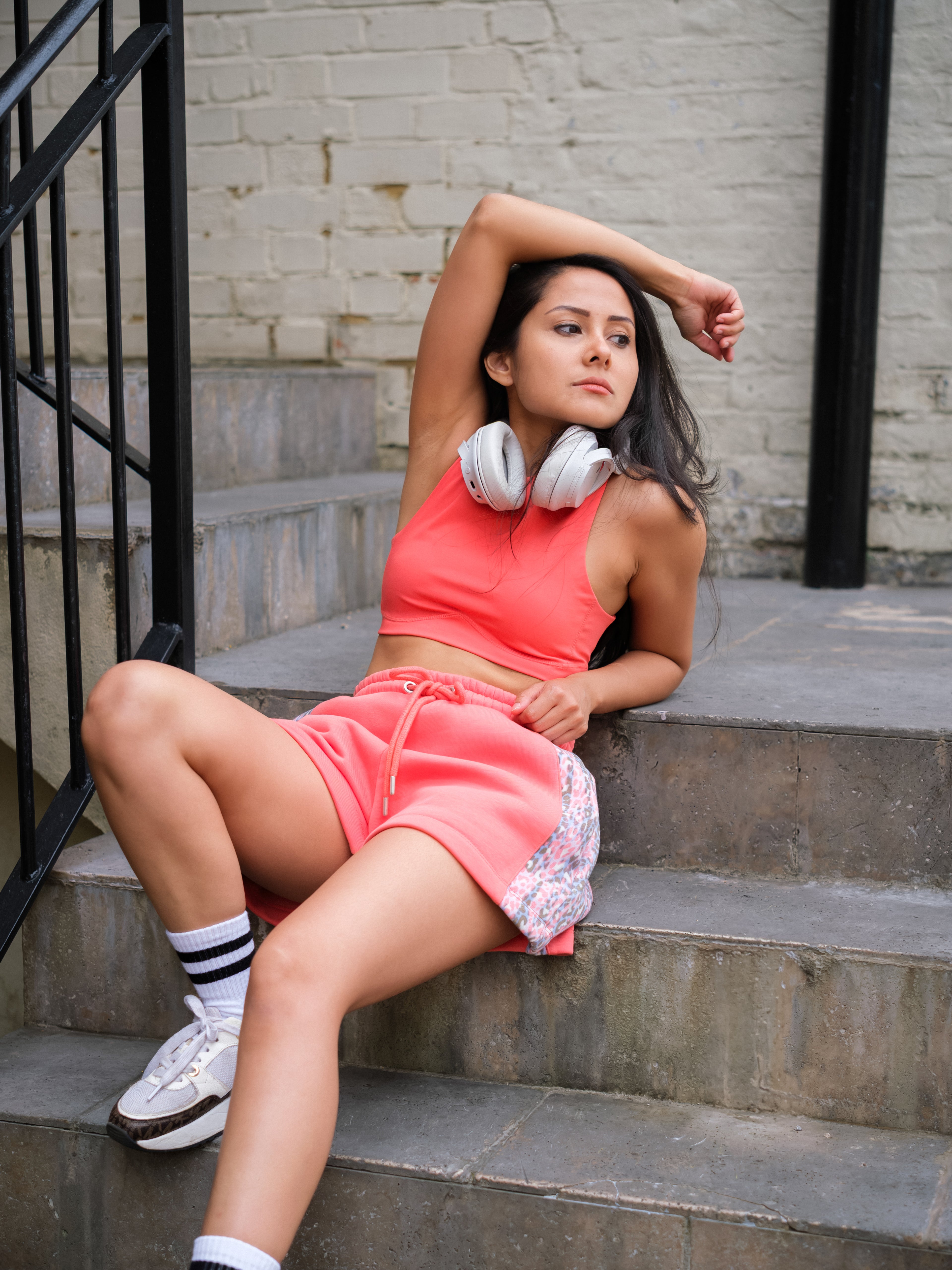 Wearing DEWEY's HAZEL Cropped Vest Top in Coral Red (Extra Petite), a woman sits relaxed on outdoor steps with headphones around her neck, pairing the athleisure look with white sneakers and striped socks.