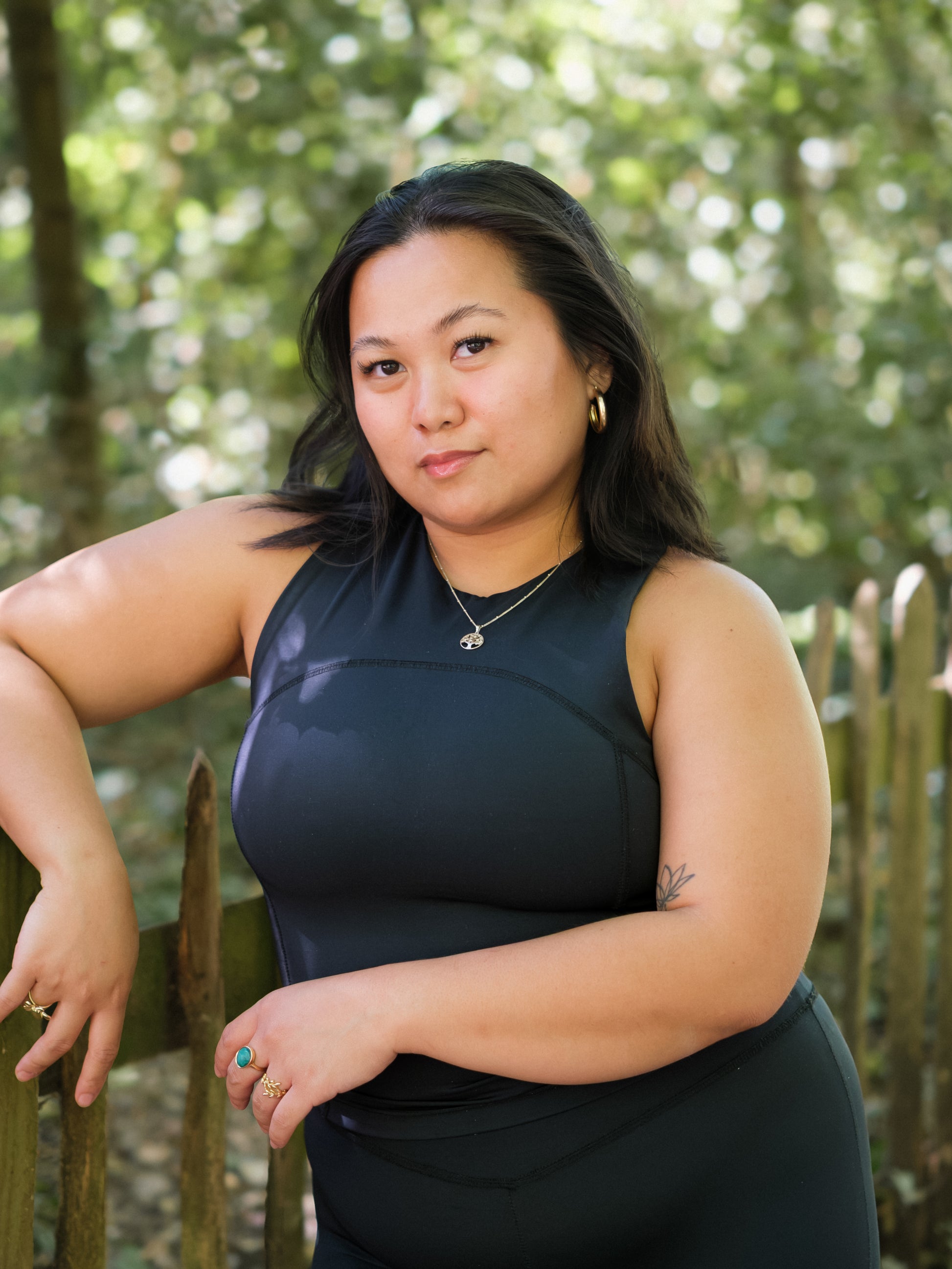 A woman with long dark hair stands outdoors by a wooden fence, wearing the DEWEY SUMMER Racerback Vest Top in Black (Extra Petite), paired with gold hoop earrings and a pendant necklace. Blurred green foliage forms the background.