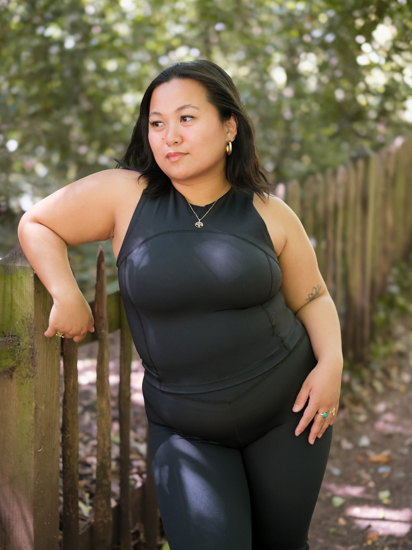 A woman with dark hair stands outdoors by a wooden fence, wearing the DEWEY SUMMER Racerback Vest Top in Black (Extra Petite). She looks to the side, surrounded by green trees and dappled sunlight.