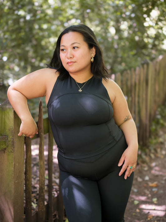 A woman with dark hair stands outdoors by a wooden fence, wearing the DEWEY SUMMER Racerback Vest Top in Black (Extra Petite). She looks to the side, surrounded by green trees and dappled sunlight.