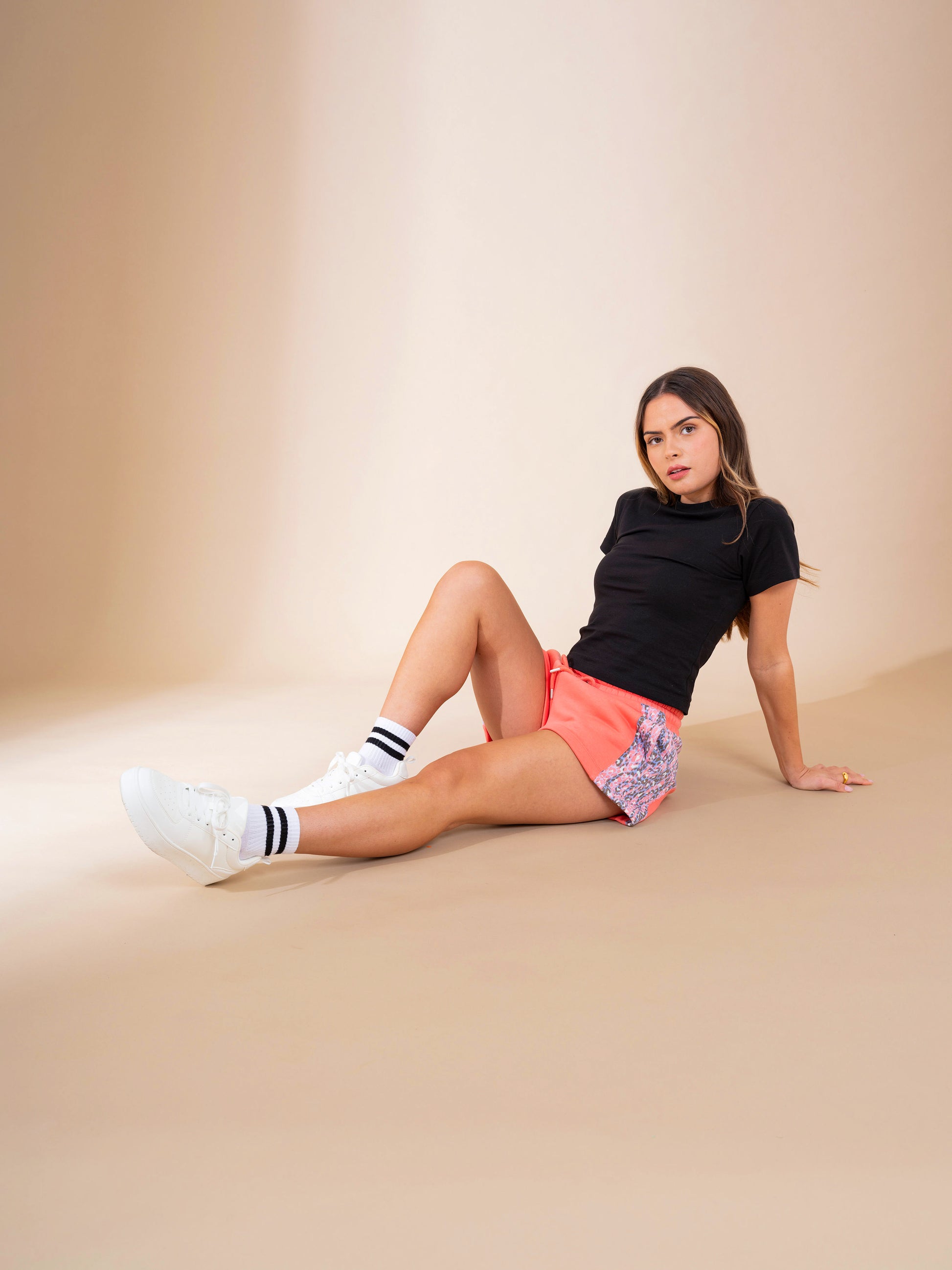 A woman sits on the floor against a beige background, wearing the DEWEY ALEX Slim Fit Short Sleeve T-Shirt Tee in black (Extra Petite). She pairs it with pink shorts, white striped socks, and white sneakers, looking directly at the camera.