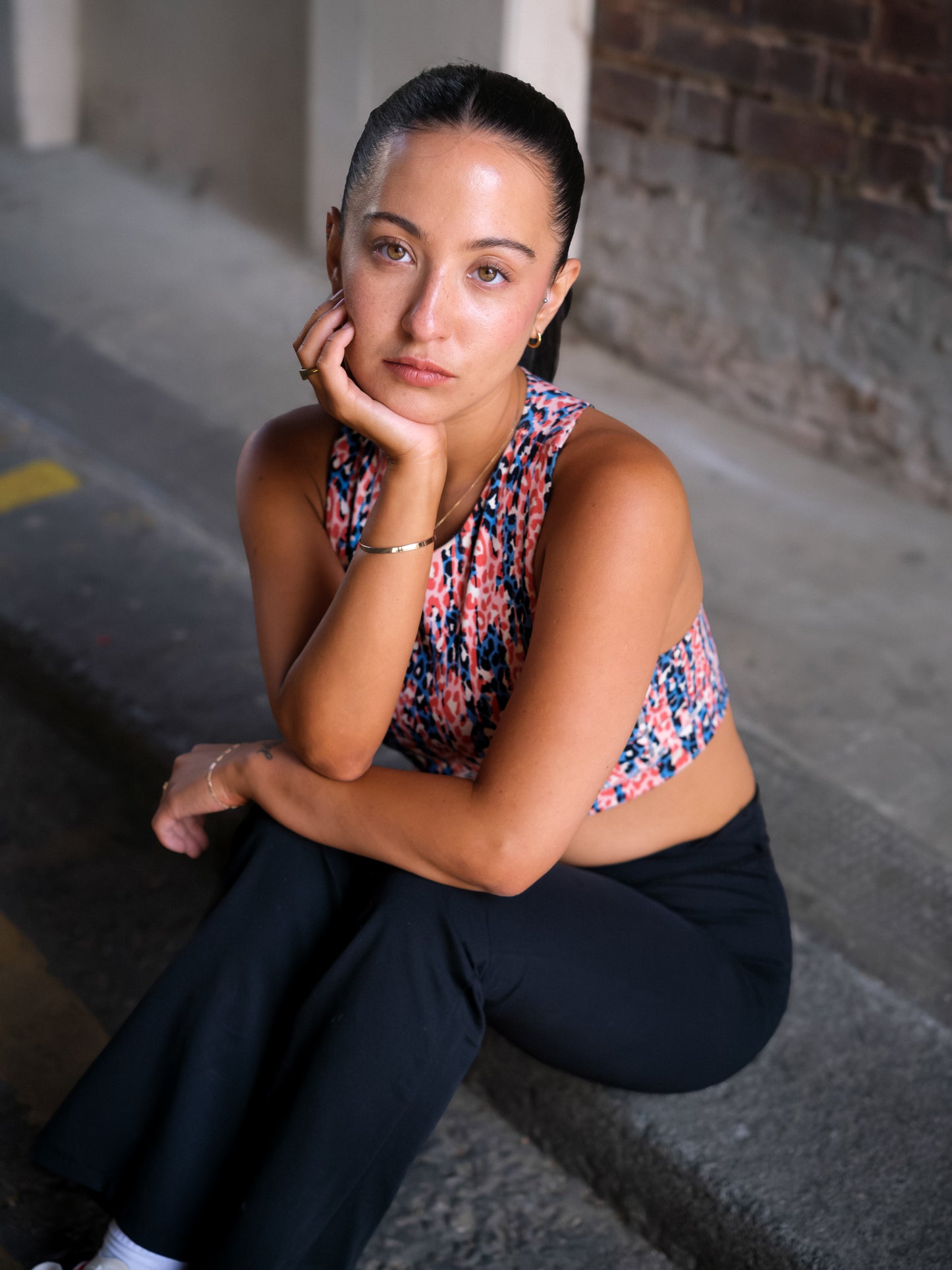 A woman with dark hair tied back sits on a curb, resting her chin on her hand. She wears the DEWEY HAZEL Cropped Vest Top (Printed, Extra Petite) and black pants, gazing thoughtfully at the camera by an urban brick wall.