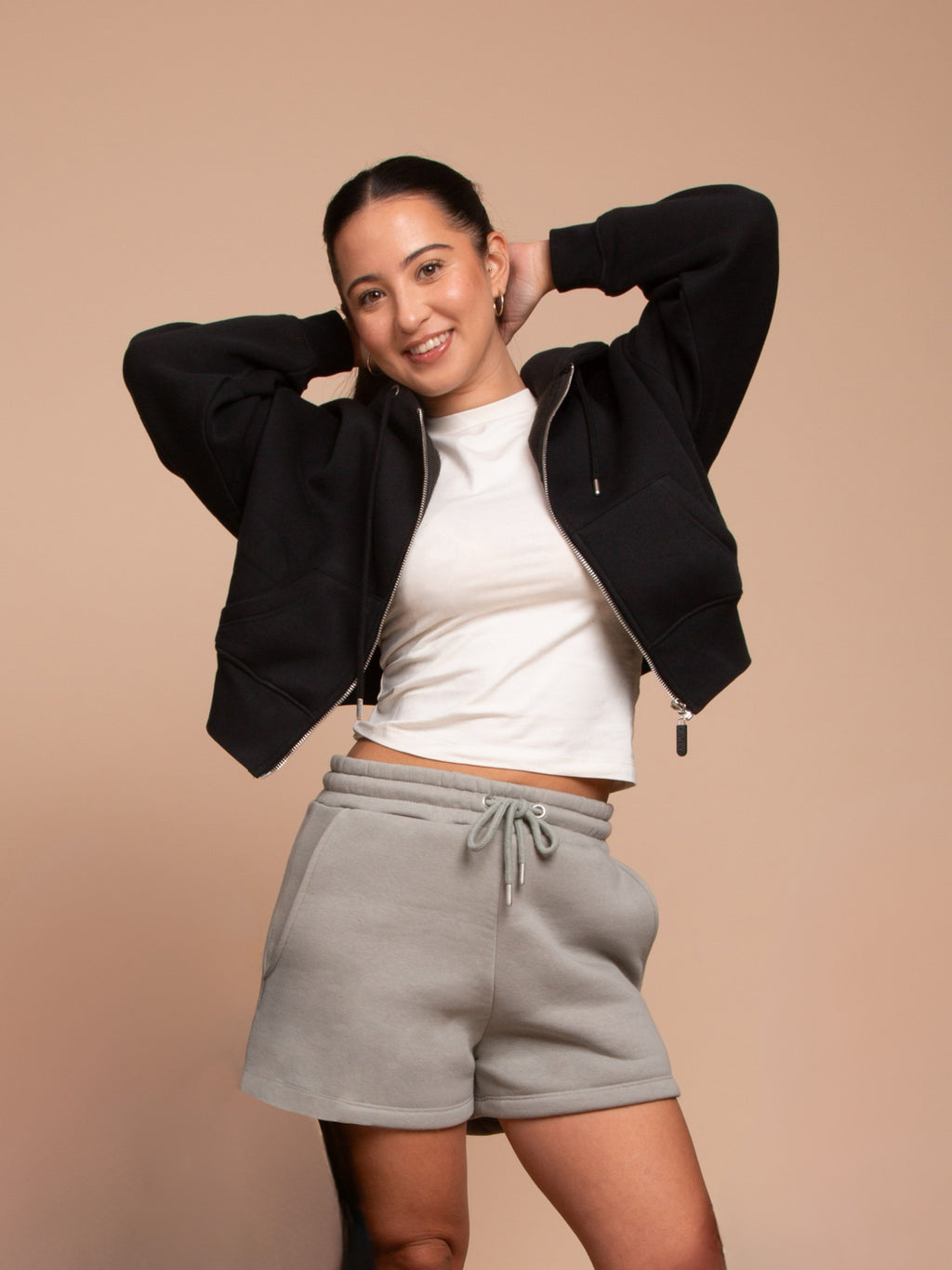 A young woman smiles with her hands behind her head, wearing the DEWEY TOBY Full Zip-Up Hoodie in black (Extra Petite), a white t-shirt, and gray drawstring shorts against a plain beige background.