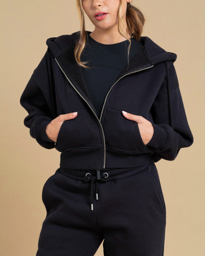 A woman wears the DEWEY TOBY Full Zip-Up Hoodie in black (Extra Petite) with matching sweatpants, standing against a beige background with her hands in the front pockets. The sleek hoodie’s zip-up style complements her partially visible face.