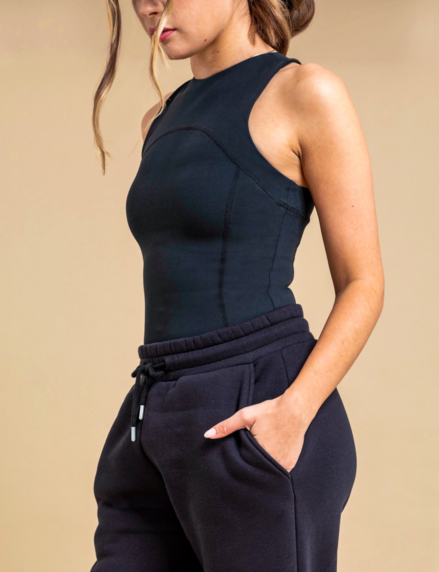 A woman wears the DEWEY SUMMER Racerback Vest Top in Black (Extra Petite) with black jogger pants, her left hand in her pocket as she stands against a beige background. Her face is partly out of frame.