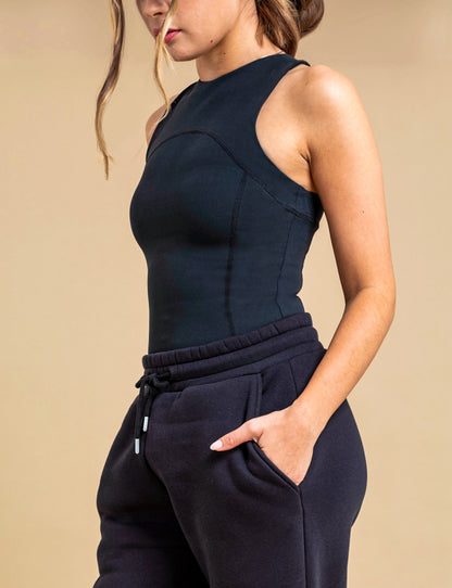 A woman wears the DEWEY SUMMER Racerback Vest Top in Black (Extra Petite) with black jogger pants, her left hand in her pocket as she stands against a beige background. Her face is partly out of frame.