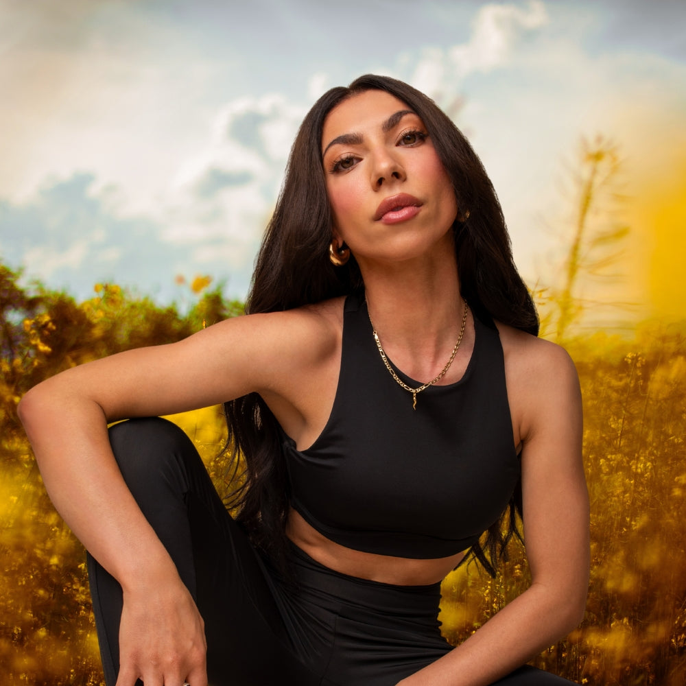 A person with long dark hair, wearing a black sleeveless crop top and leggings, poses outdoors in front of a field with yellow flowers and a cloudy sky.