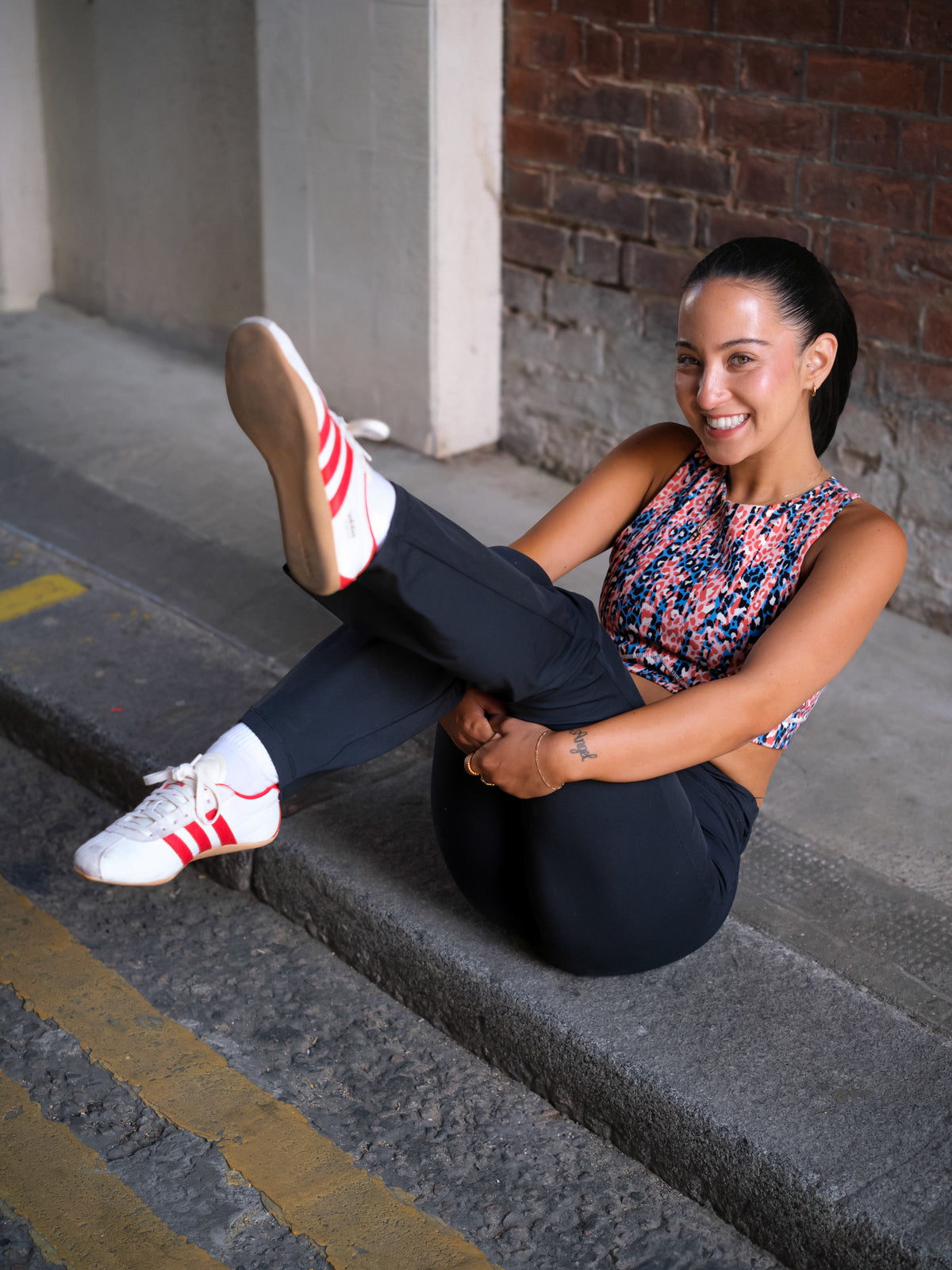 A woman in athleisure sits on a curb, smiling and playfully holding up one leg. She wears a colorful DEWEY HAZEL Cropped Vest Top (Printed, Extra Petite), black pants, white socks, and white sneakers with red stripes.