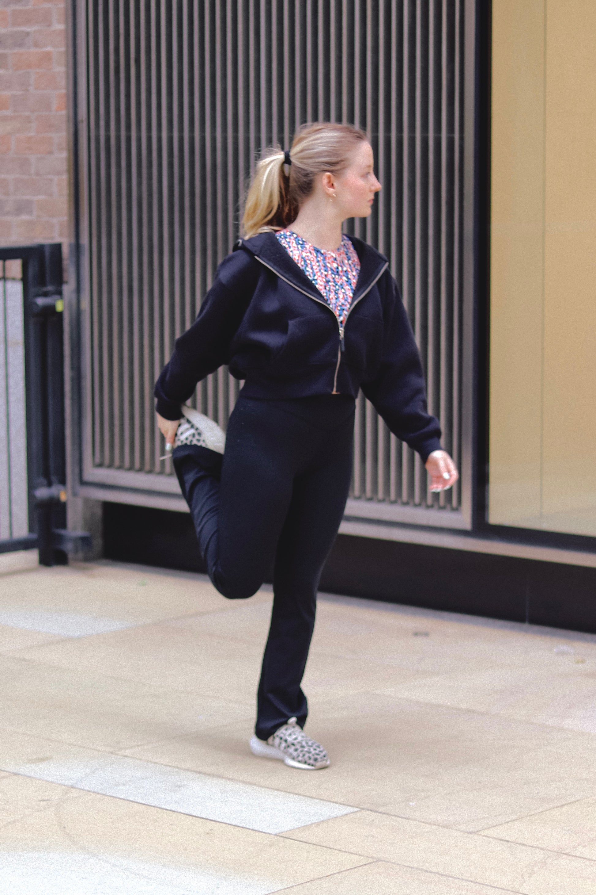 A woman in a DEWEY TOBY Full Zip-Up Hoodie (Black, Extra Petite) stretches her leg, holding her ankle behind her while standing on one foot and gazing to the side in an urban outdoor setting.