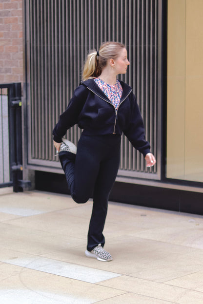 A woman in a DEWEY TOBY Full Zip-Up Hoodie (Black, Extra Petite) stretches her leg, holding her ankle behind her while standing on one foot and gazing to the side in an urban outdoor setting.