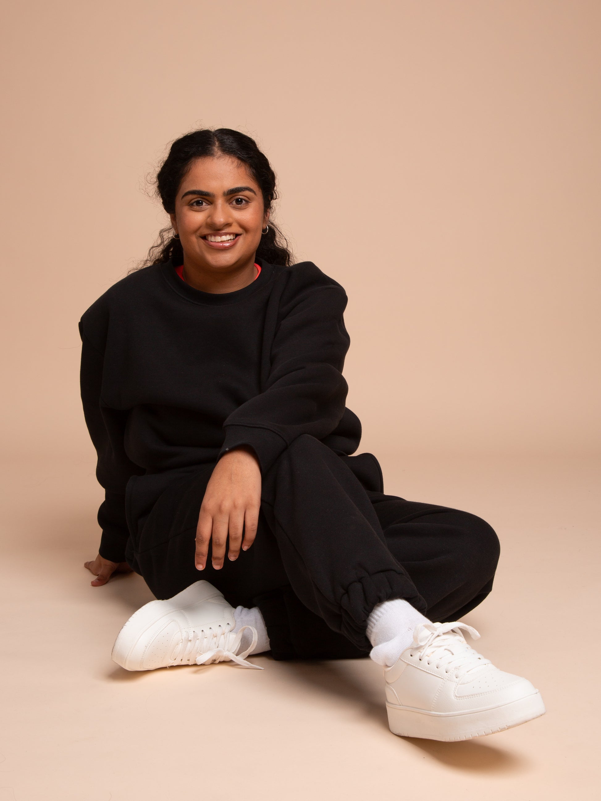 A person with dark hair in a ponytail smiles at the camera while sitting on the floor against a beige background, wearing a relaxed fit DEWEY BAILEY Crewneck Sweatshirt Jumper (Black, Extra Petite), sweatpants, white socks, and chunky sneakers.