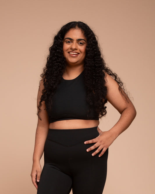 A woman with long, curly hair smiles while wearing the DEWEY HAZEL Cropped Vest Top in black (Extra Petite), posing against a plain beige background.