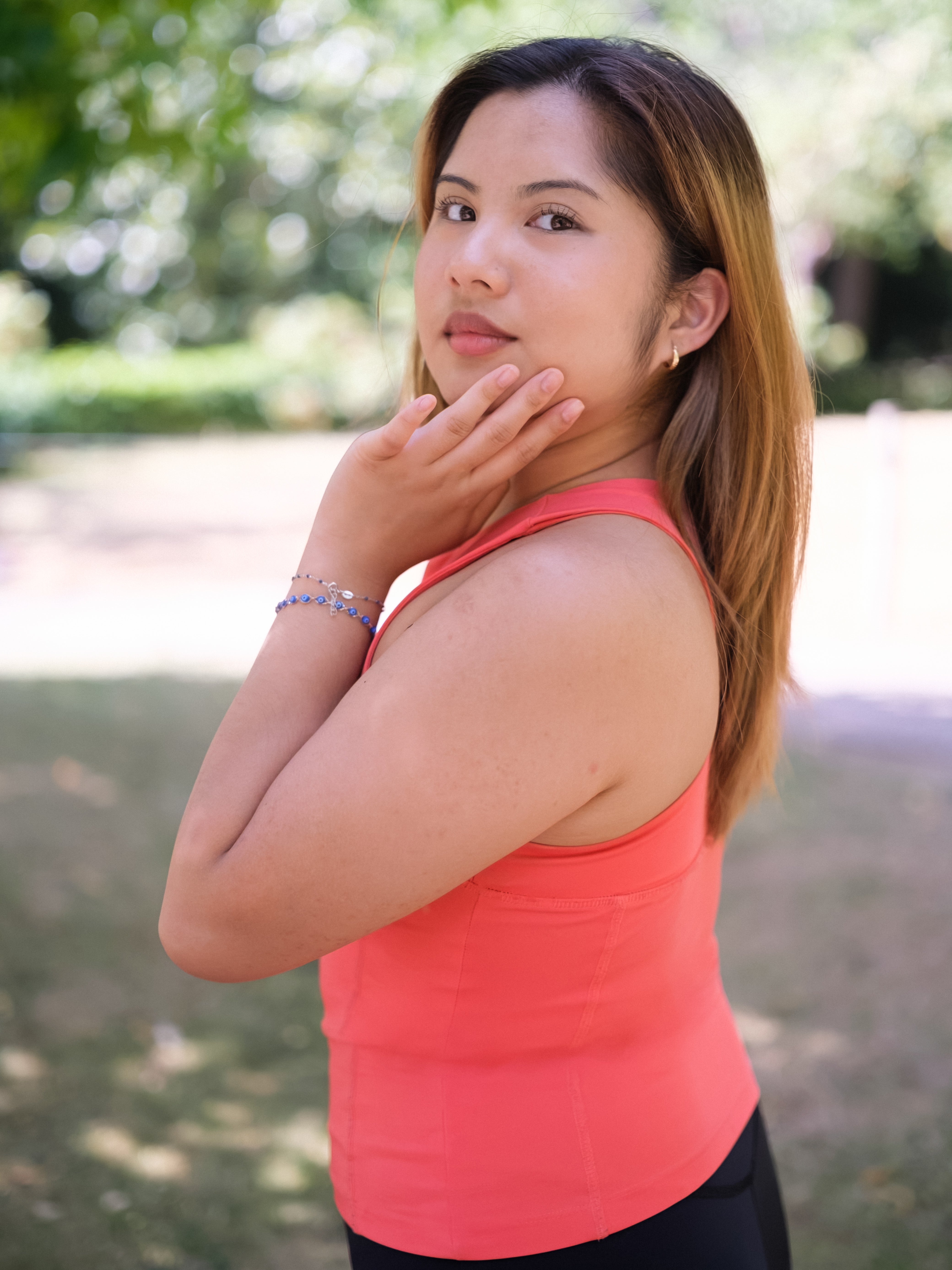 A woman with long hair wears the DEWEY SUMMER Racerback Vest Top in Coral Red (Extra Petite) outdoors, gently touching her face and looking at the camera with greenery blurred in the background.