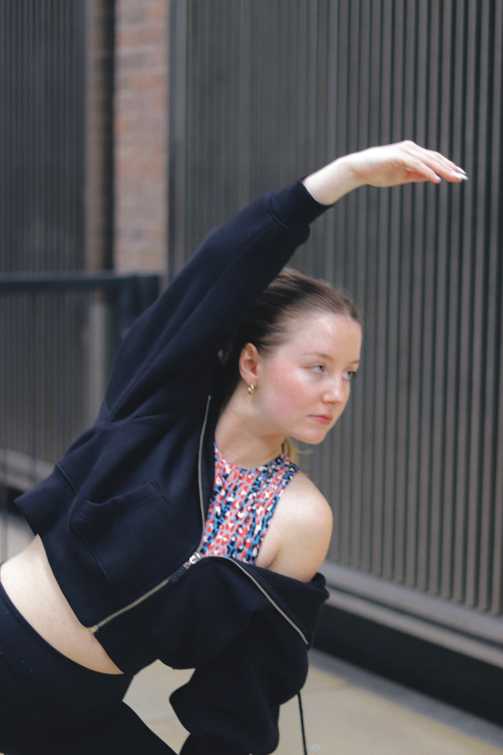 Wearing the DEWEY TOBY Full Zip-Up Hoodie (Black, Extra Petite), a woman stretches outdoors in front of a building with vertical metal bars, raising one arm overhead and tilting her torso, her hair pulled back in focus.