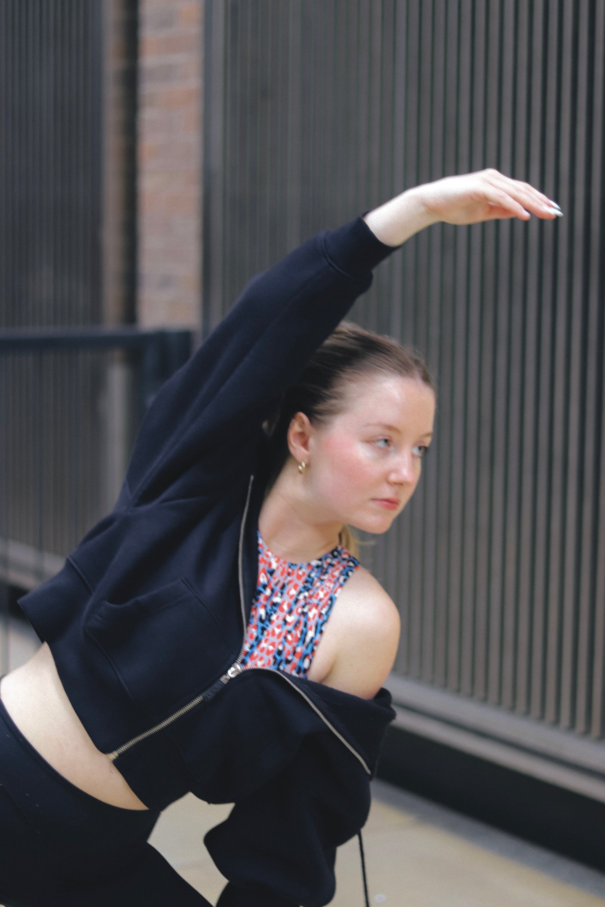 Wearing the DEWEY TOBY Full Zip-Up Hoodie (Black, Extra Petite), a woman stretches outdoors in front of a building with vertical metal bars, raising one arm overhead and tilting her torso, her hair pulled back in focus.