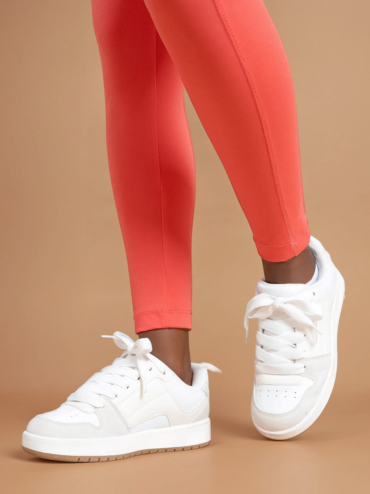 A person wears DEWEY IRIS High Waist Leggings in Coral Red (Extra Petite) and white sneakers, standing on a tan surface with one foot slightly raised to show the side and front of the shoes.