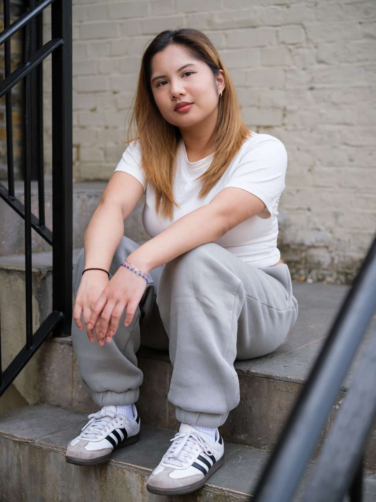 A young woman with straight, light brown hair sits outdoors in a white t-shirt and DEWEY RILEY Sweatpants Joggers (Grey, Extra Petite), modeling effortless petite loungewear with her arms resting on her knees.