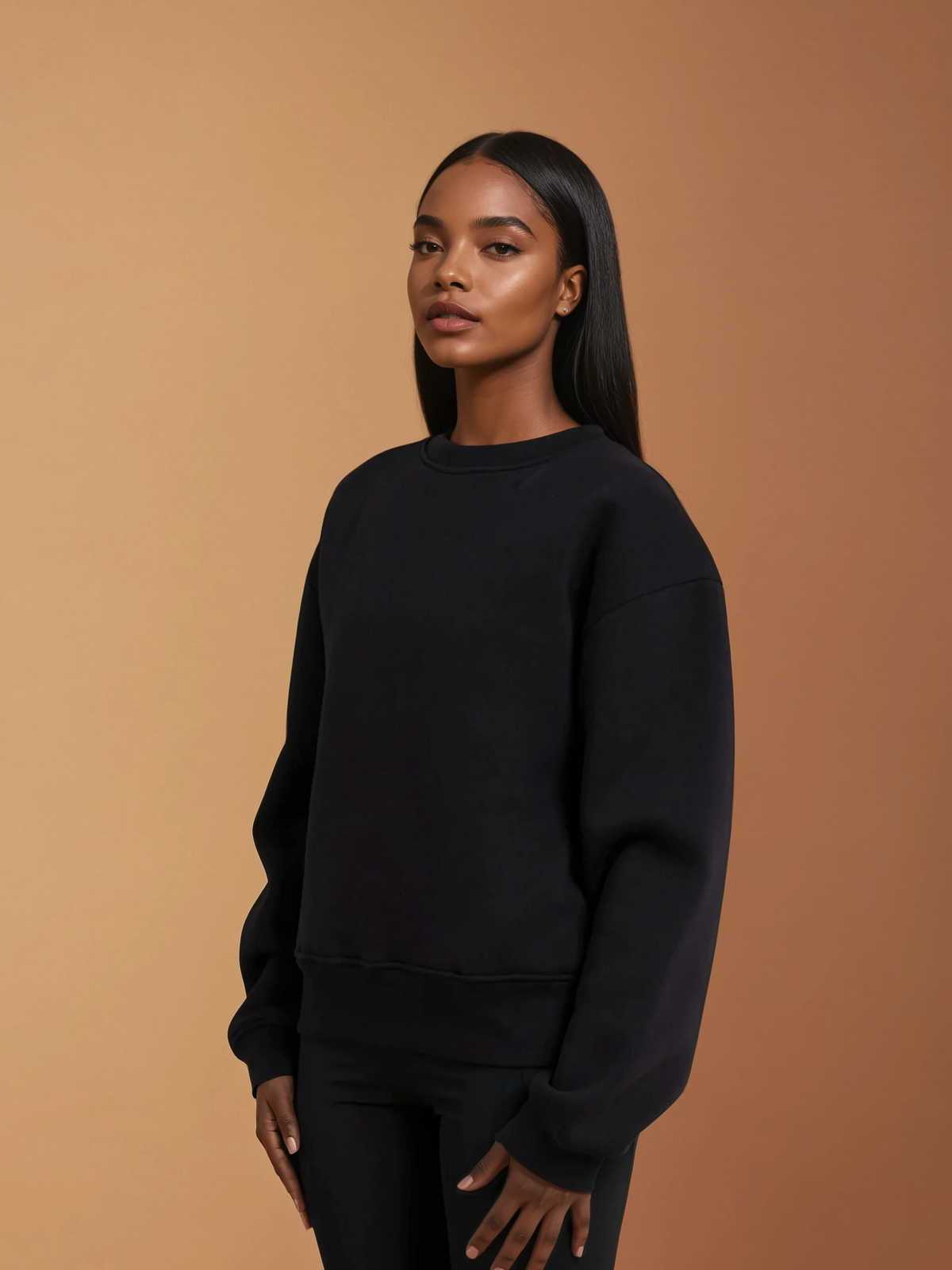 A woman with long straight hair wears the DEWEY BAILEY Crewneck Sweatshirt Jumper in Black (Extra Petite), paired with black pants, standing against a beige background and looking slightly to the side with a neutral expression.