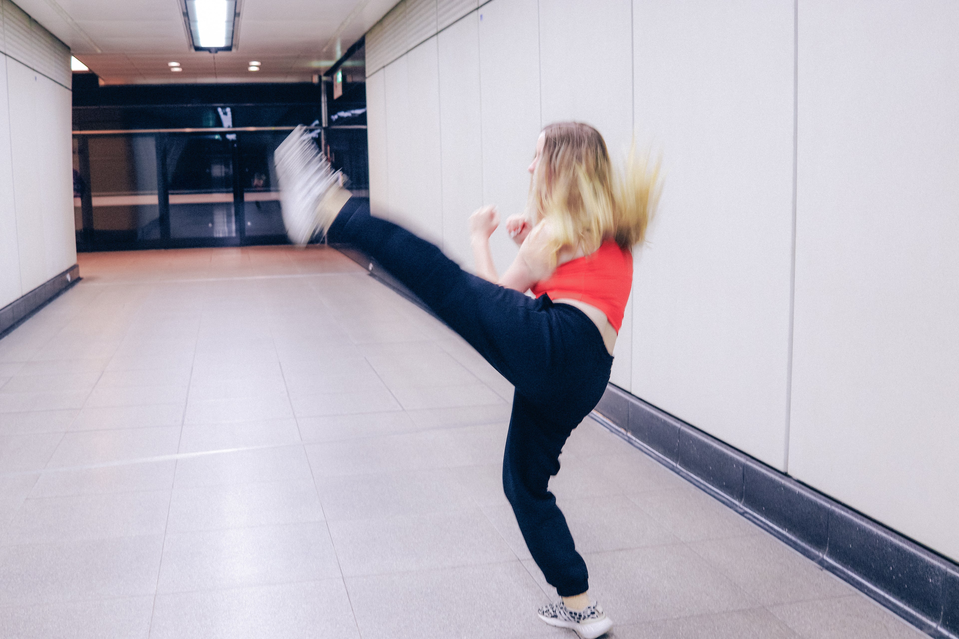 A person with long blonde hair, wearing a red sleeveless top and black pants, performs a high kick in an empty, well-lit hallway with tiled floors and white walls.